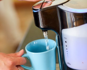 A kettle on the right with a light blue mug underneath being filled with water. There is a hand holding the mug.