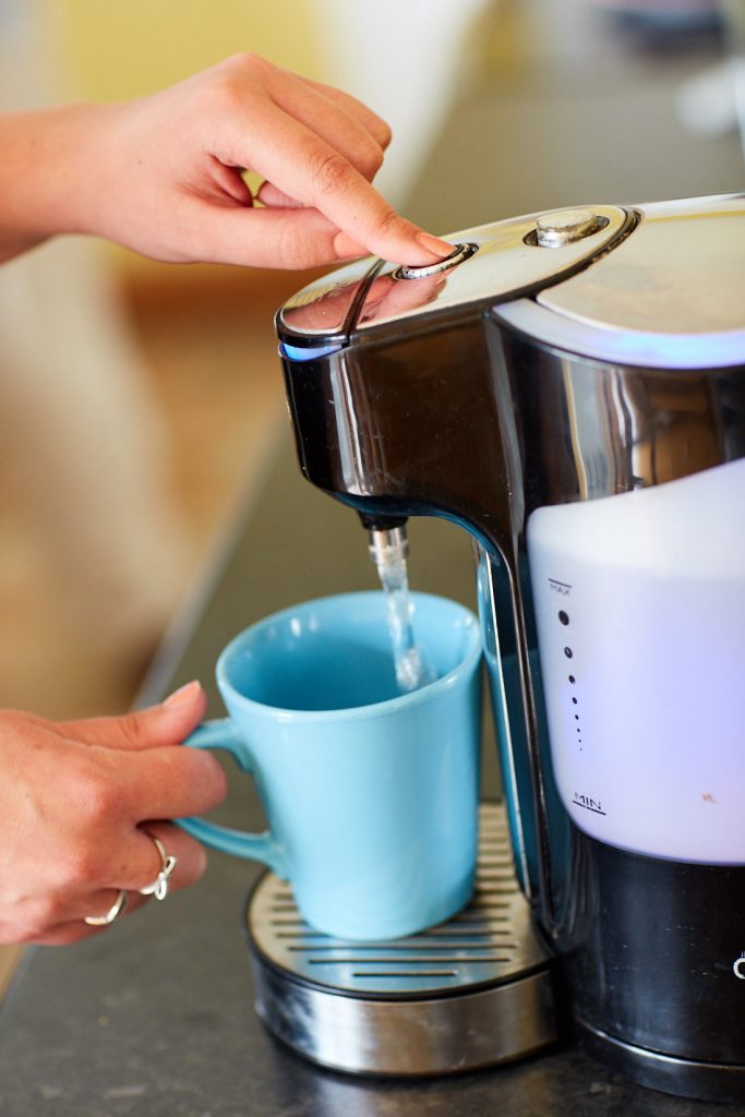A kettle on the right with a light blue mug underneath being filled with water. There is a hand holding the mug.