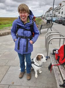 A woman with short hair, a light blue coat and rucksack smiling next to a white and browndog Dexter.