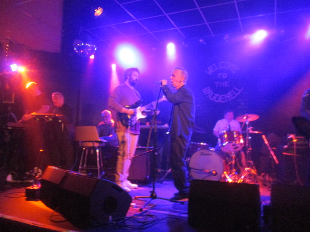 A photo looking onto a stage with a man with a guitar in the centre foreground alongside a man holding onto a microphone stand. In the background there is a man on the drums and off to the right there are three other men, one is playing the keyboard.
