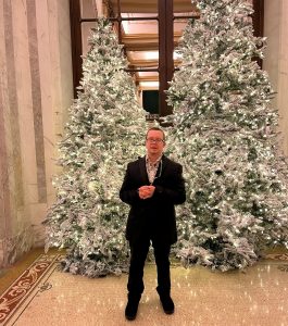 A man wearing a black suit standing in front of two Christmas trees.