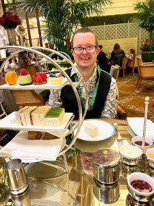 A man in a patterned shirt and black waistcoat sat at a table. The table is centred with a large afternoon tea stand full of cakes, desserts and sandwiches.