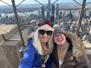 A blonde woman in a blue coat and a man in a brown coat smiling in front of a view of the New York skyline.