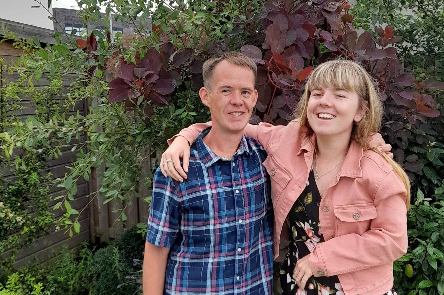 Two people stand in a garden with their arms round each other.