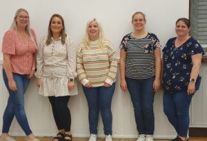 Five women standing in a  line against a white wall.