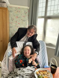 A woman sat in bed with a man in a suit standing behind her.