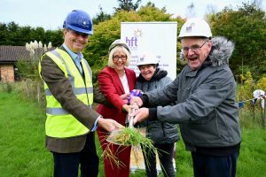 From left to right - a man in a high-vis, a woman in a red suit, a woman in a grey coat and a man in a grey coat all wearing hard hats and holding a shovel.
