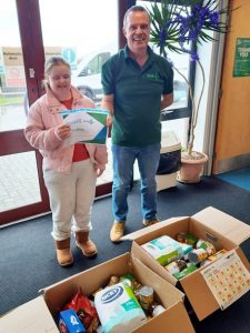 Rachel with her certificate and a foodbank worker, standing behind two boxes of donations