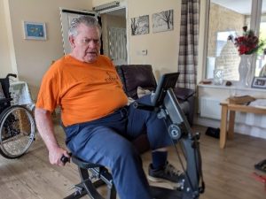 Gordon, wearing an orange shirt with the FitBees logo, on his exercise bike
