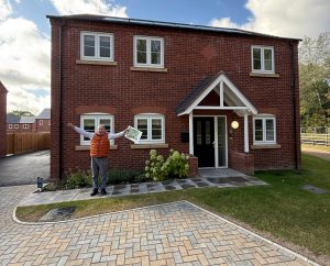 Simon stands with outstretched arms outside his new house. It's clad in red brick and has white window frames and an awning over the front door.