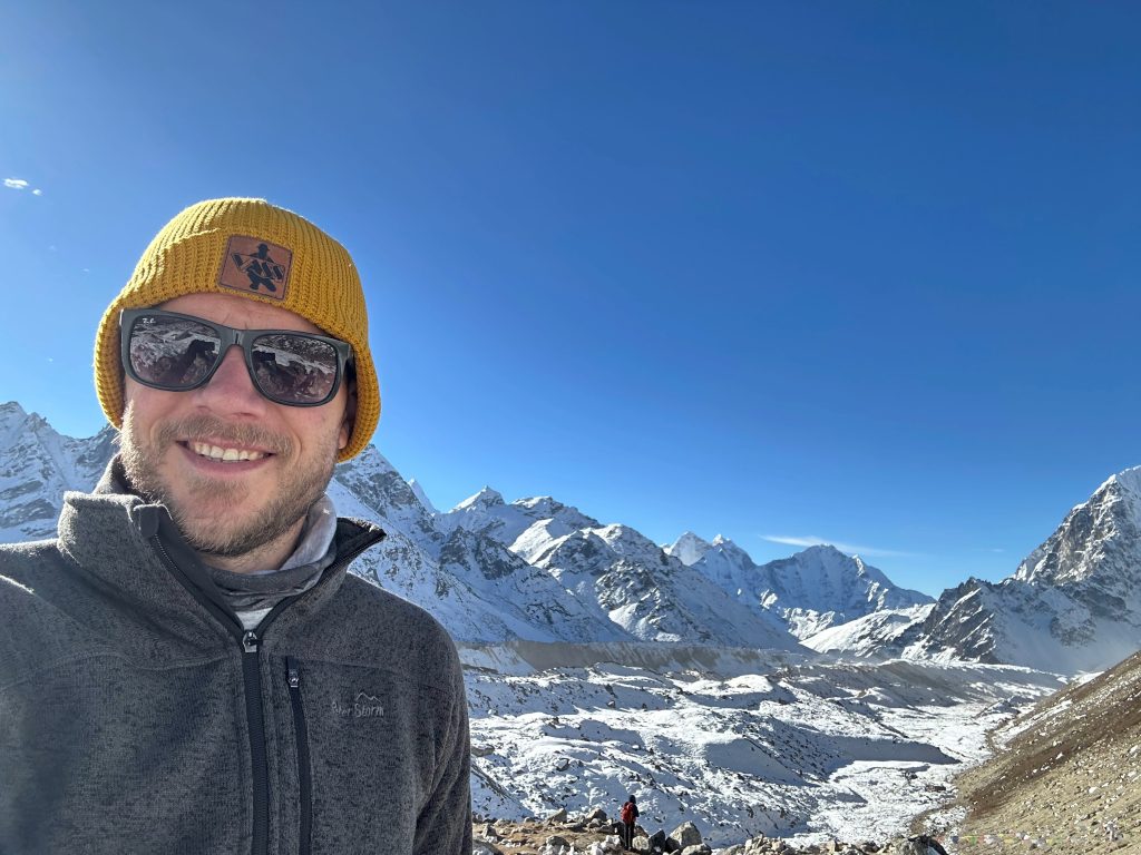 Ian takes a selfie with snowy mountains in the background and a big blue sky. 