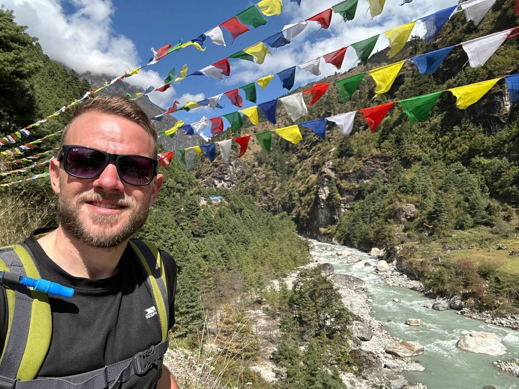 Ian crosses a mountain river surrounded by evergreen forest. There's colourful cloth bunting along the trail. 