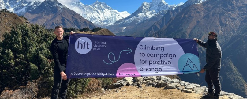 Ian and guide Budda hold up a large banner that reads "HFT learning disability allies. Climbing to campaign for positive change." In the background is the snow-capped mountain range.