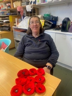 Anne sits at a table. On the table are nine of her knitted red poppies.