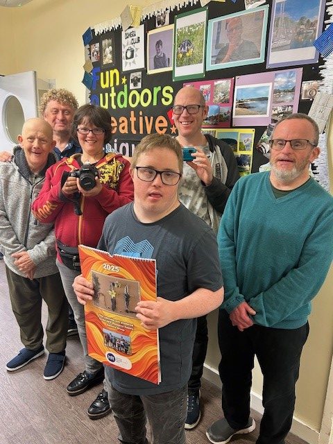 The photography group smile for a photo. Some are holding cameras whilst a man in the middle holds last year's calendar.