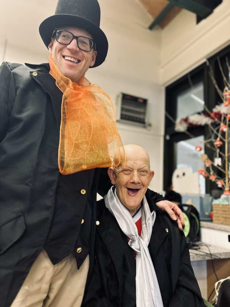 Two of the actors smiling. Colin is standing and has his arm around Darren who's sat in a chair. 