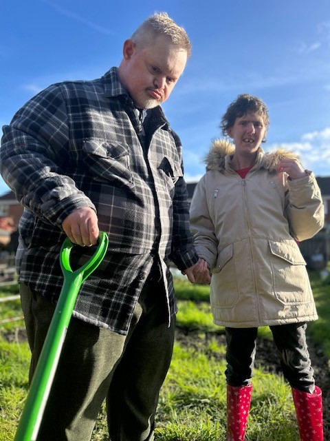 Two of the gardening volunteers hold hands at the allotment. One is also holding a green spade handle.