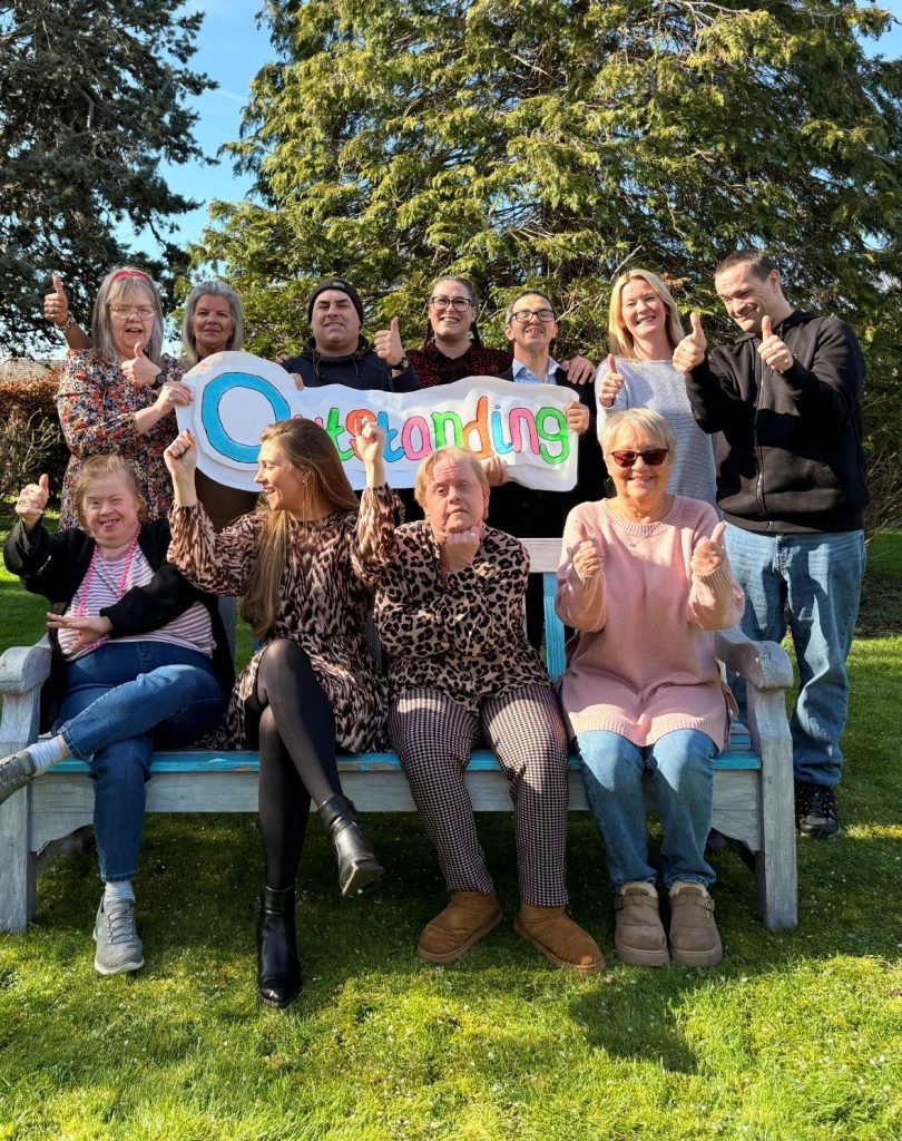 Four colleagues and people we support sit on a wooden bench in a sunny garden. Seven more stand behind them, holding a homemade banner that says 'outstanding;' in colourful letters. They're all smiling and holding their thumbs up.