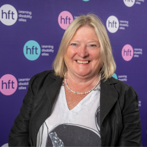 Mairi smiles against a dark purple backdrop. She's got shoulder-length blonde hair and is wearing a black and white top and a black jacket.